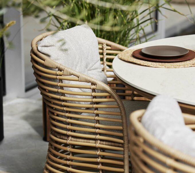 Der runde Aspect Esstisch von Cane-line mit Fossil-Keramikplatte im Travertine look mit Basket Sesseln hellbraunen Wove-Kissen.