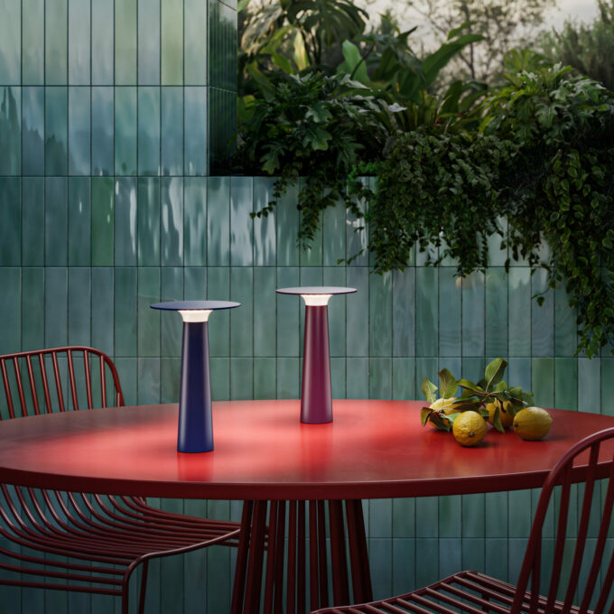 Zwei Lix Akku-Leuchten stehen auf einem roten Gartentisch. Die Lampen haben die Farbe aubergine und midnight blue.